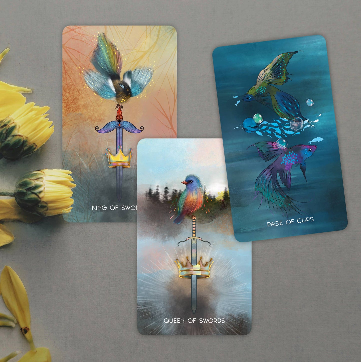 Three tarot cards with colorful illustrations on a gray surface with yellow flowers.