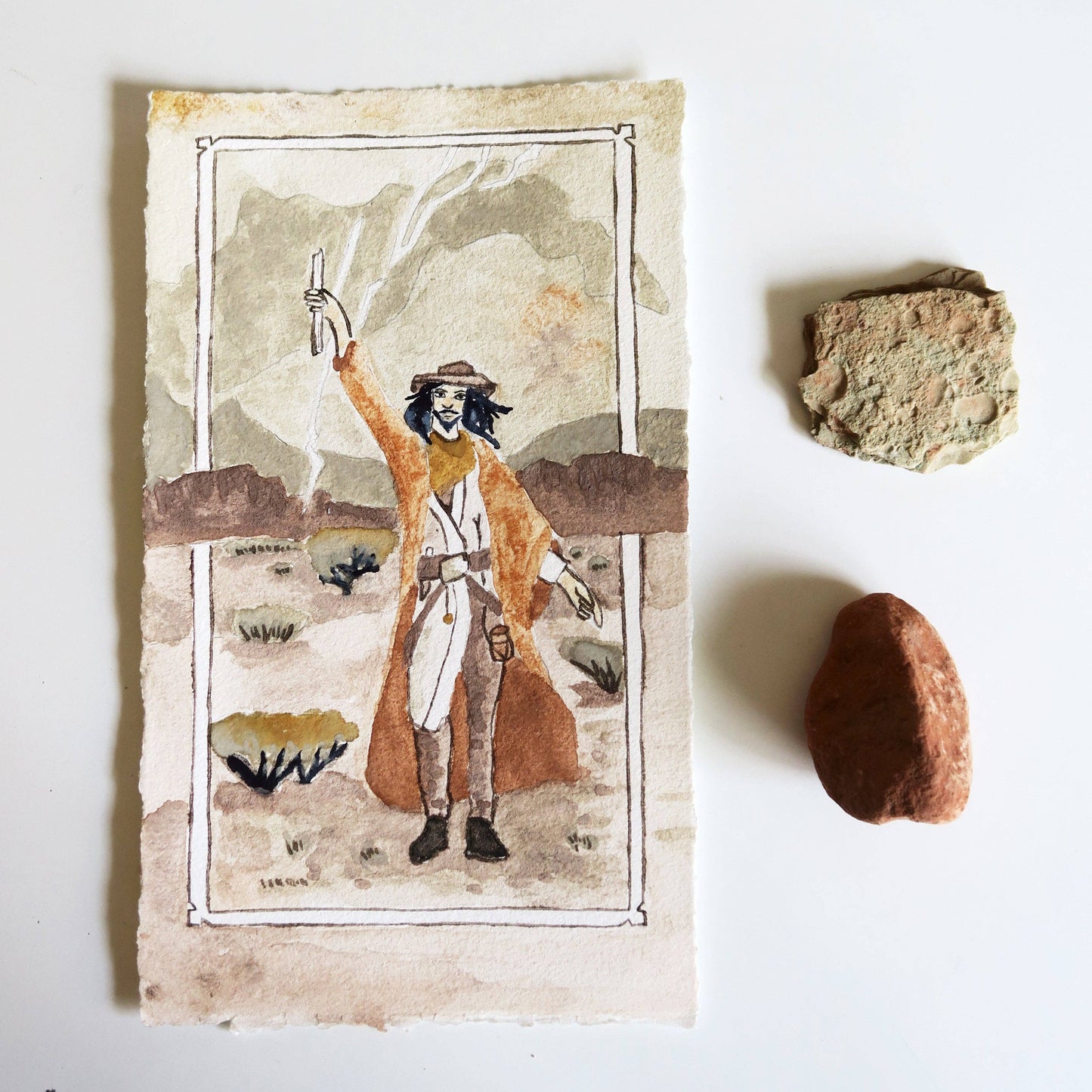 A square handmade paper art print featuring a hand-painted western scene with a figure holding a rifle, displayed on a white surface next to two small natural stones.