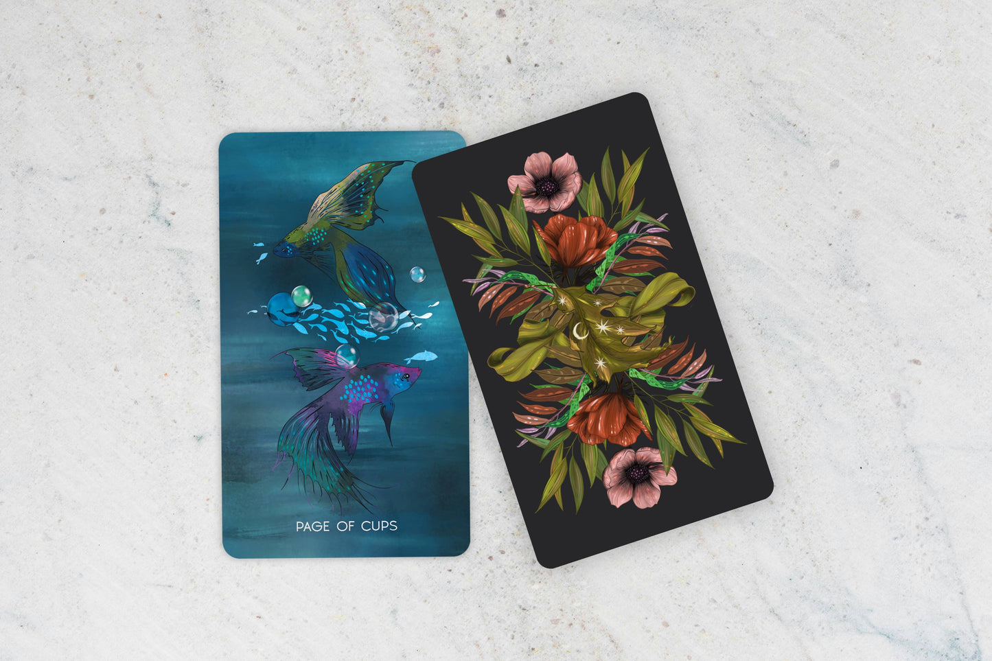 Two tarot cards on a textured surface, one with fish and the other with floral design.