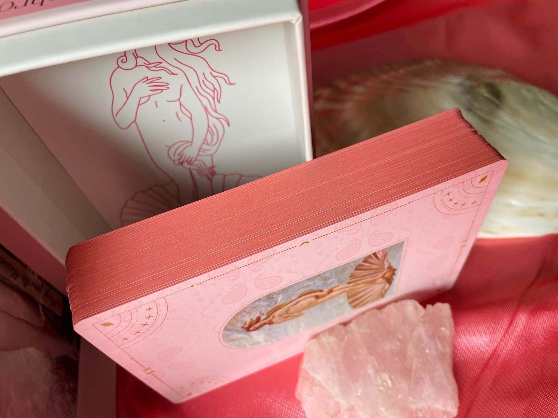 Showing limited edition details of romantic pink edges for the cards of the Musing of Aphrodite oracle card deck supported by a chunk of rose Quartz