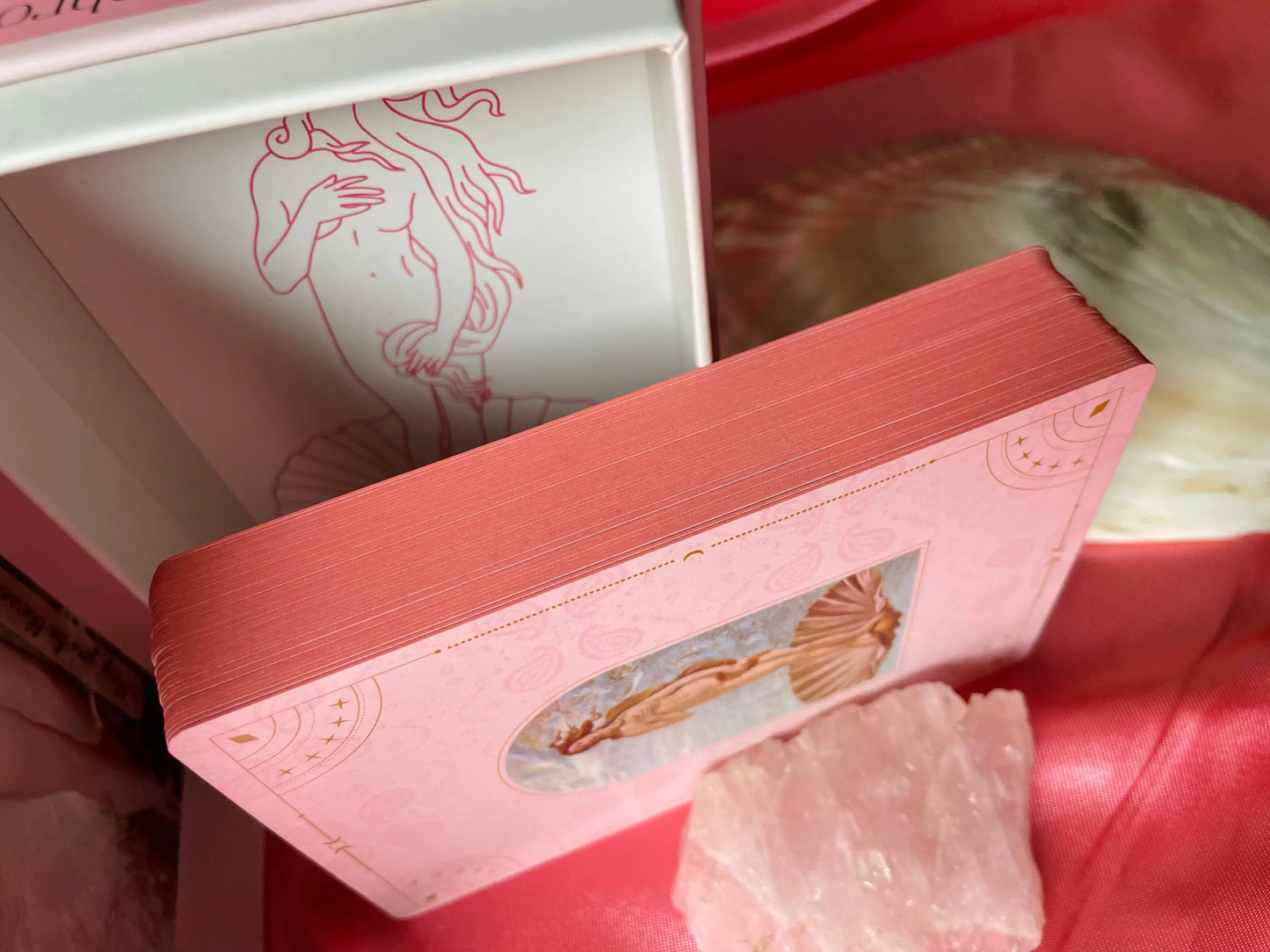 Showing limited edition details of romantic pink edges for the cards of the Musing of Aphrodite oracle card deck supported by a chunk of rose Quartz