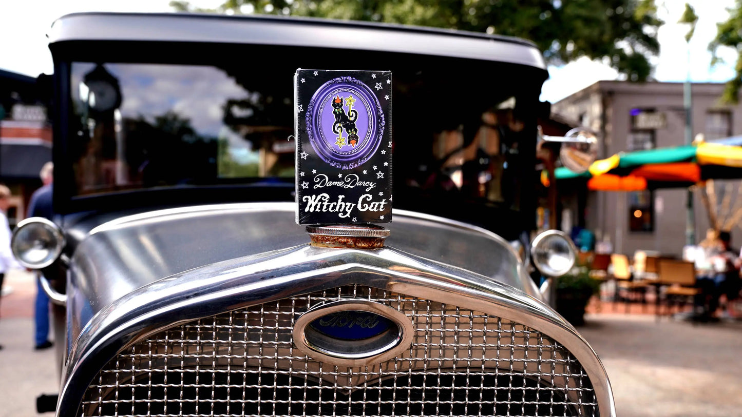Vintage car with a 'Witchy Cat' sign on the grill in an outdoor setting