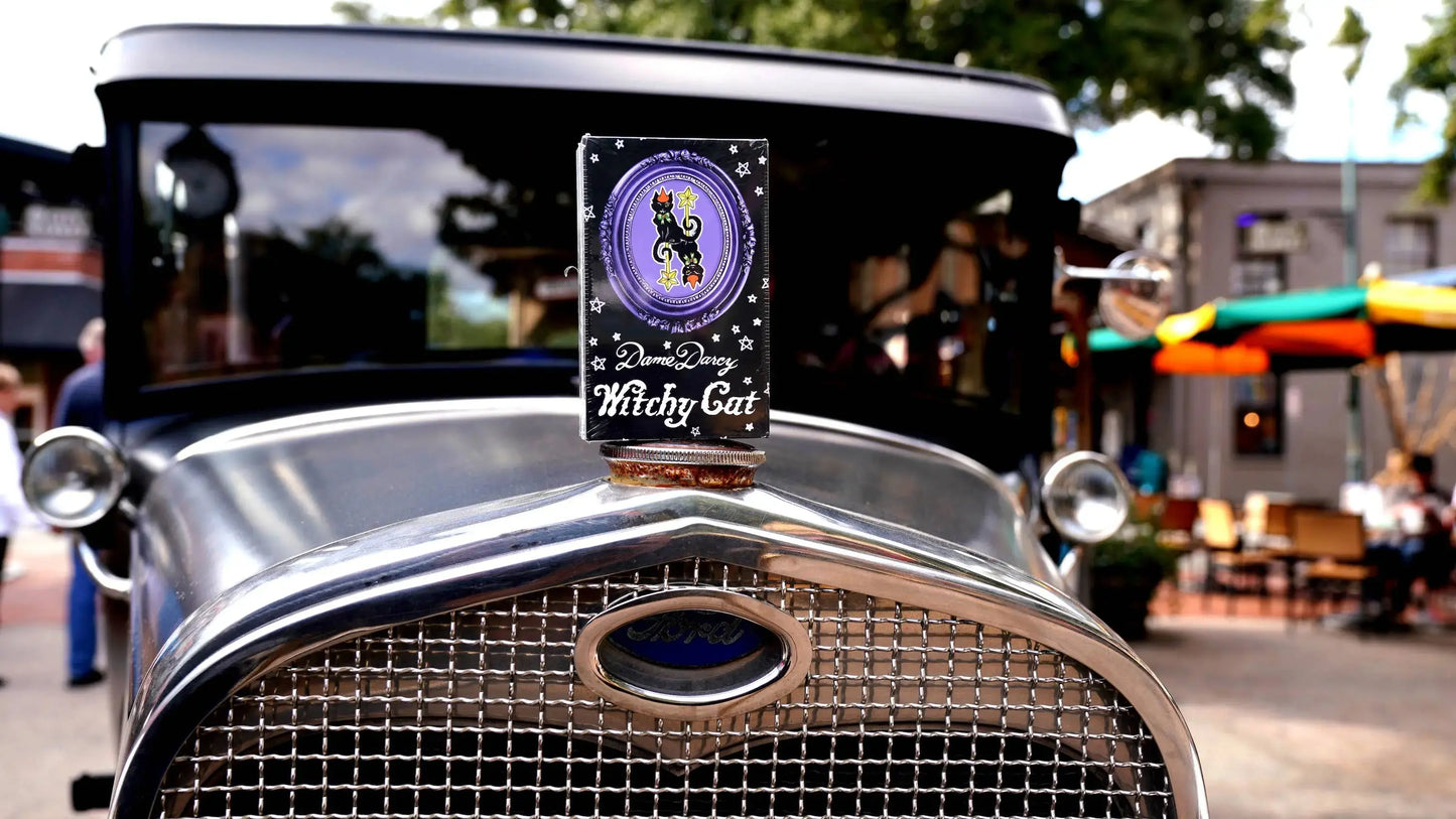 Vintage car with a 'Witchy Cat' sign on the grill in an outdoor setting