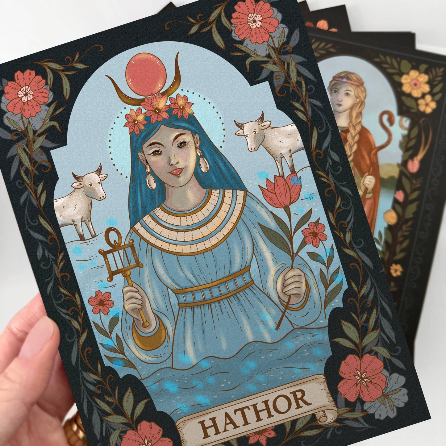 Card featuring an illustration of Hathor with floral and animal elements, held by a hand.
