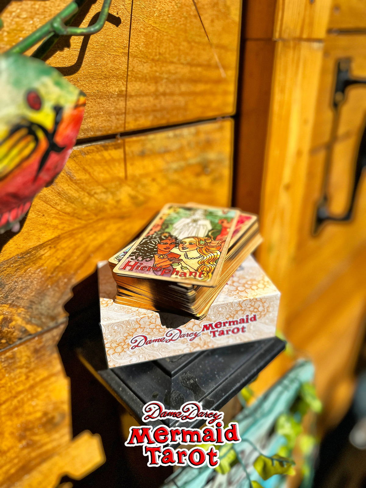 A deck of Dream Dance Mermaid Tarot cards displayed on top of its illustrated box, set against a rustic wooden background with a decorative parrot figurine partially visible on the left.