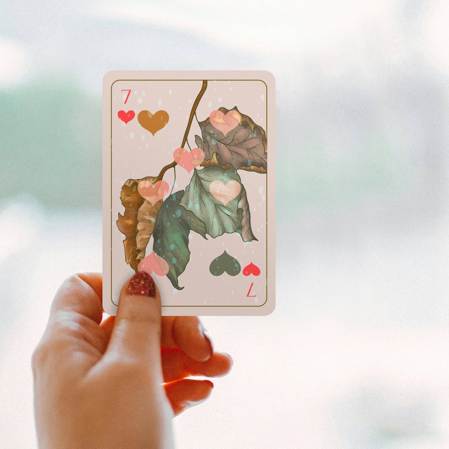 Hand holding a playing card with leaf design and heart symbols on a blurred background