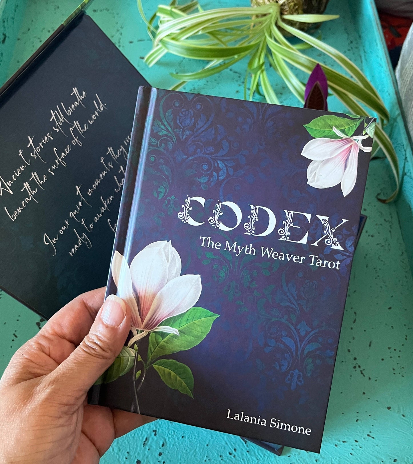 Codex The Myth Weaver Tarot book held by a hand with a decorative cover featuring floral designs on a teal surface.