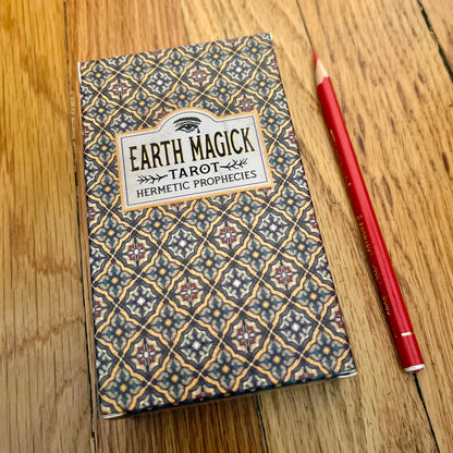 Earth Magick Tarot deck box with a geometric patterned cover, placed on a wooden tabletop next to a red pencil.