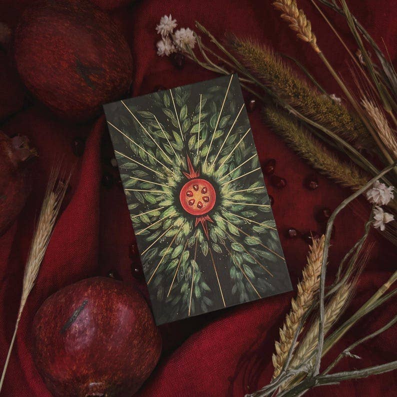 A tarot card deck box featuring a green leafy sunburst design with a red pomegranate illustration in the center, styled in a flat lay with red fabric, pomegranates, dried wheat, and small white flowers. Asphodelon Tarot by Asphodelon Studio featuring hand-painted Greek mythology artwork, gilded edges, premium matte cards and a deluxe gold foil presentation box.