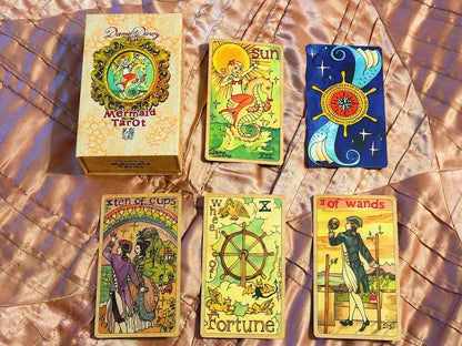 Collection of tarot cards and their box on a textured surface Illustrated mermaid-themed tarot cards from Dame Darcy Mermaid Tarot Ultra Gold Edition