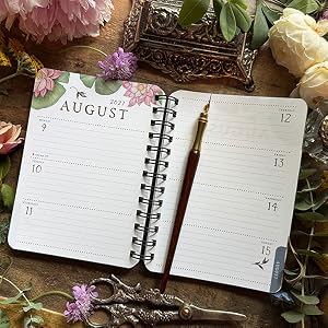 An open spiral-bound planner or journal spread showing the August weekly layout, styled with flowers, scissors, and a decorative object on a rustic wooden surface.