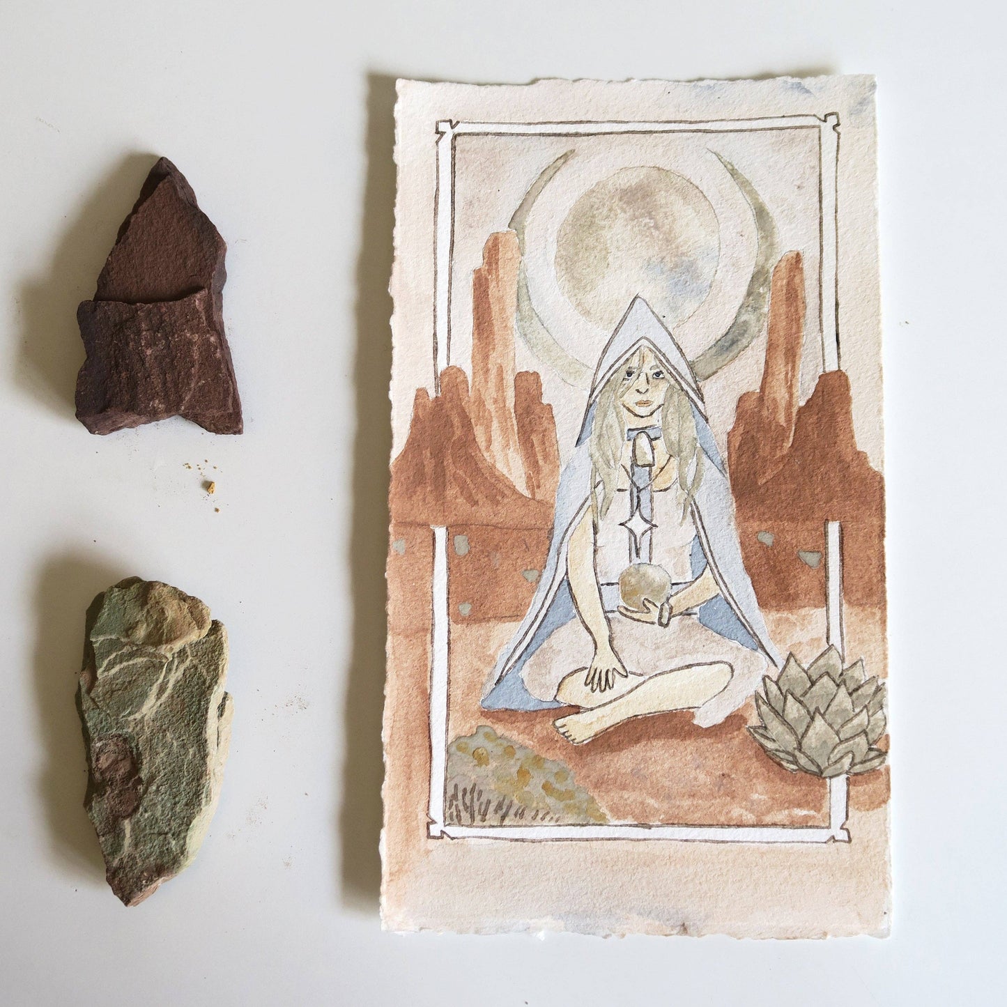A watercolor tarot-style card featuring a seated hooded figure in a desert landscape with a moon and rock formations, displayed on a white surface beside three small natural stones.