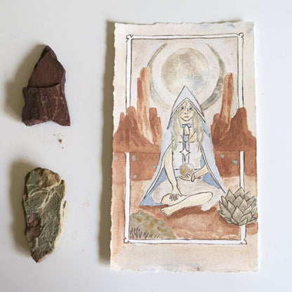 A watercolor tarot-style card featuring a seated hooded figure in a desert landscape with a moon and rock formations, displayed on a white surface beside three small natural stones.