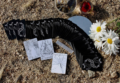 Set of black tarot cards with white designs on a textured surface with flowers. DIVINA Tarot Deck by Mary Elizabeth Evans featuring elegant monochrome pen and ink artwork with celestial symbolism and a 78-card indie tarot deck.