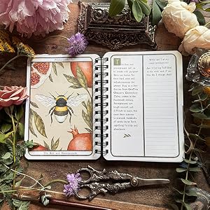 An open spiral-bound notebook displayed in a styled flat-lay arrangement with flowers, scissors, and decorative props on a rustic wooden surface. The left page features an illustration of a bumblebee with flowers and fruit, while the right page shows lined writing space with text columns.