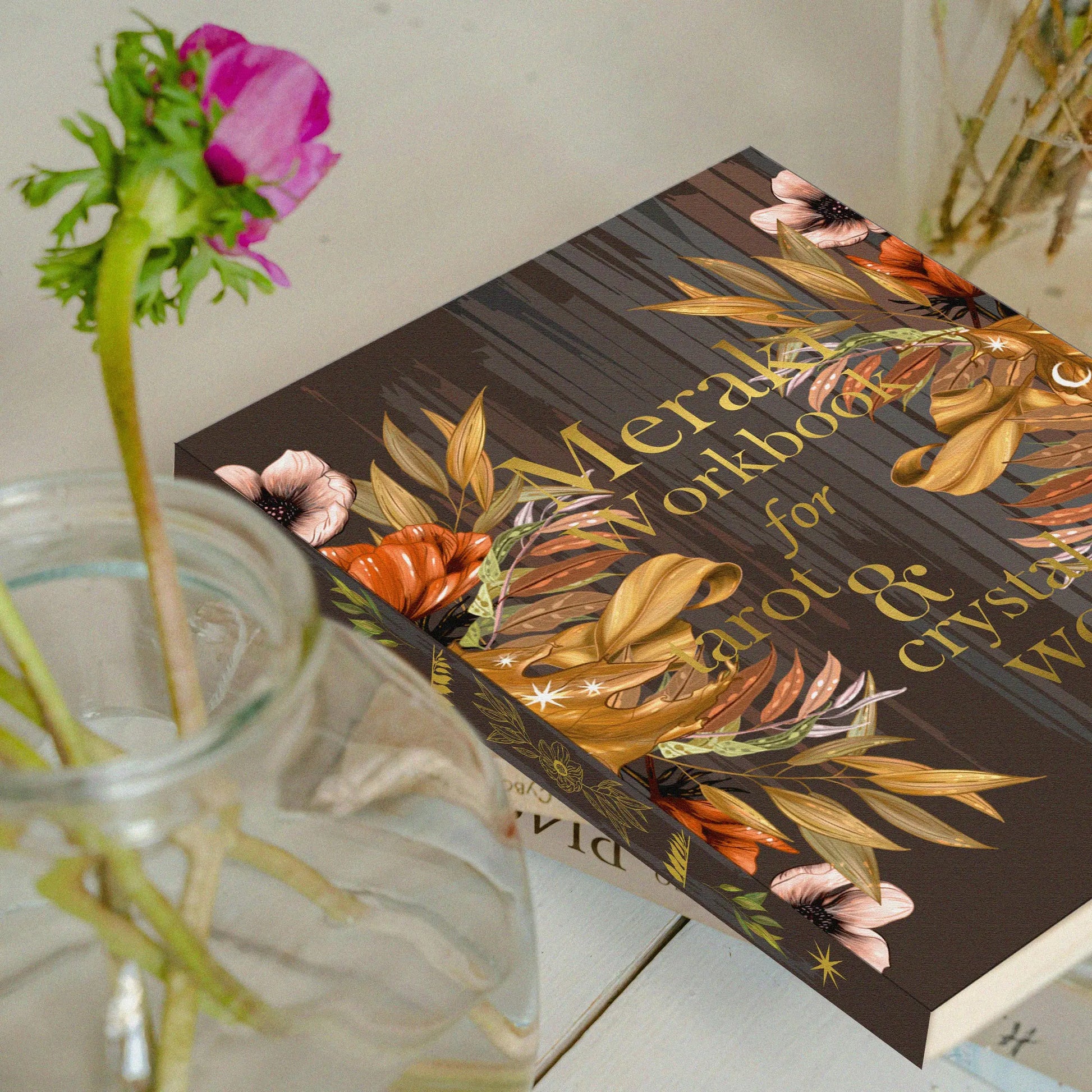 Floral-themed workbook on a surface with a glass container and flowers. Meraki workbook for tarot and crystal work 