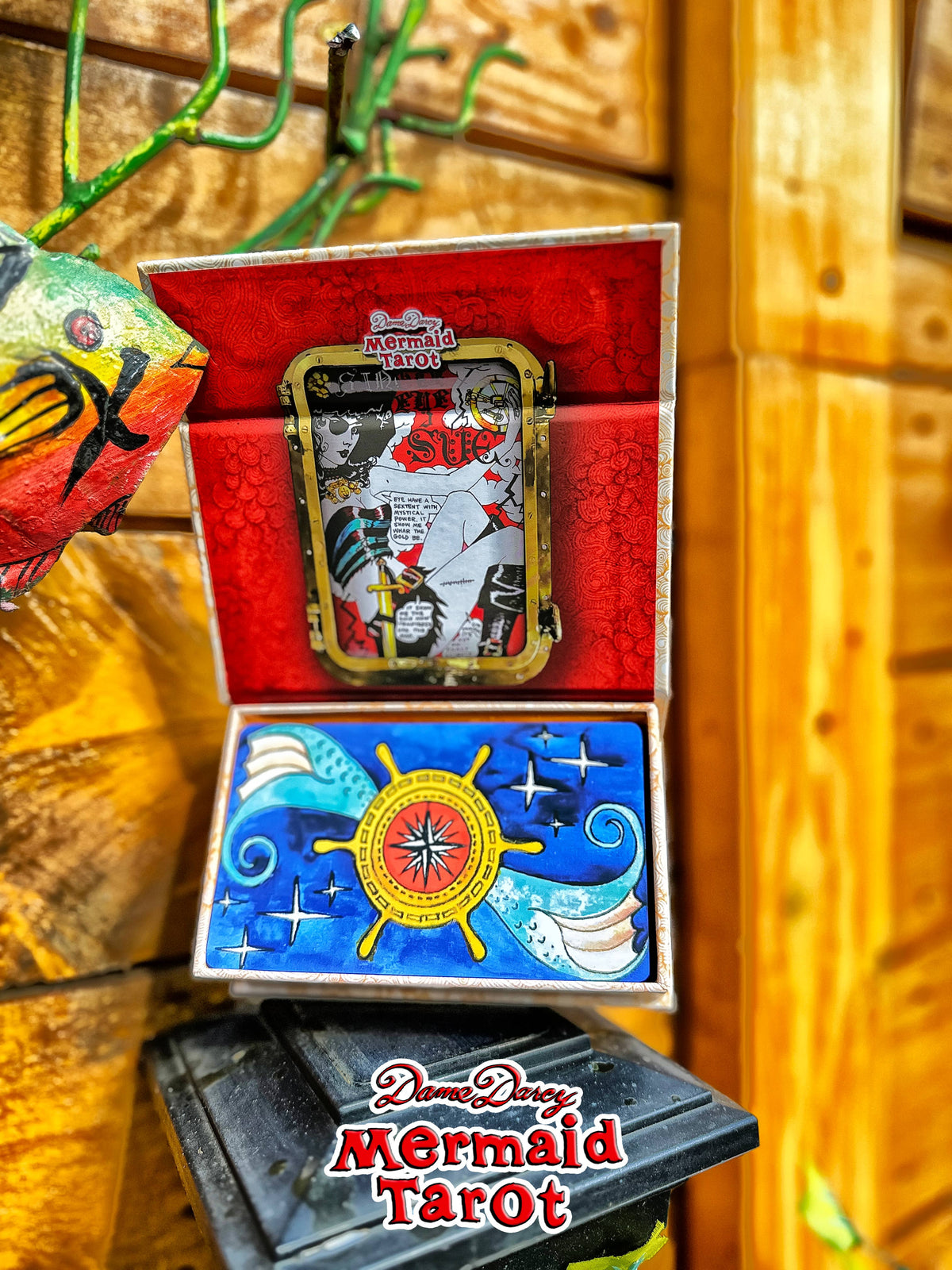 An open box of Dreamy Dreams Mermaid Tarot cards displayed against a wooden background. The top half shows the red interior with a framed mermaid card, and the bottom half shows the blue card back with a ship's wheel design.
