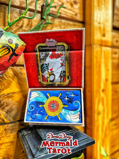 An open box of Dreamy Dreams Mermaid Tarot cards displayed against a wooden background. The top half shows the red interior with a framed mermaid card, and the bottom half shows the blue card back with a ship's wheel design.