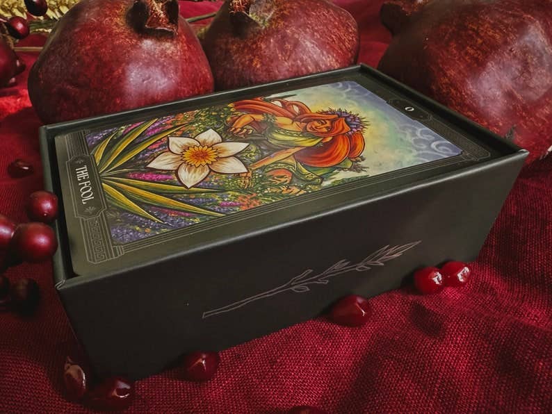 A tarot deck box labeled 'THE FOX' sits on a red fabric surface with pomegranates around it. The box lid shows a colorful illustrated card with a woman and floral artwork. Asphodelon Tarot by Asphodelon Studio featuring hand-painted Greek mythology artwork, gilded edges, premium matte cards and a deluxe gold foil presentation box.