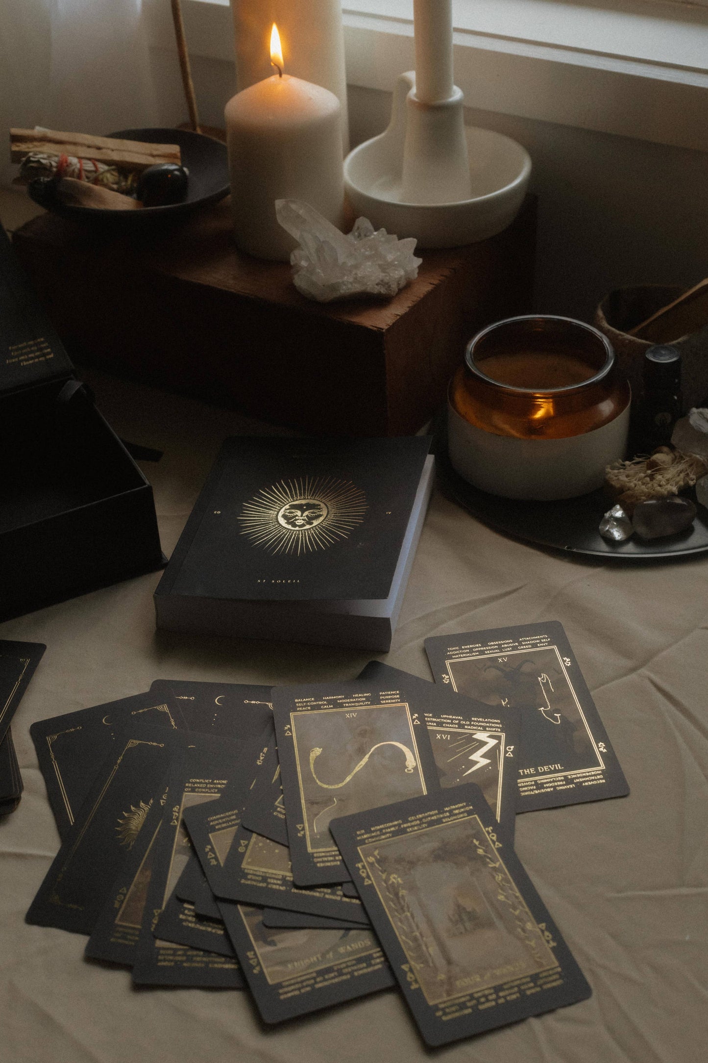 A tarot card deck and several cards are arranged on a draped fabric surface in a styled indoor setting with candles, crystals, and small ritual objects. The box and cards have a dark charcoal/black background with ornate gold illustrations.