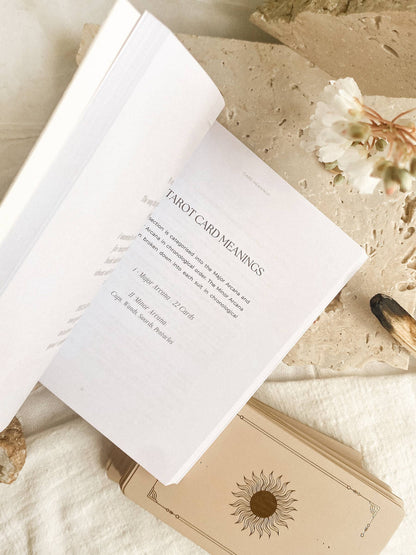 An open book or booklet showing a page titled 'TAROT CARD MEANINGS' laid on a light fabric surface, with a beige tarot card deck box underneath and dried flowers nearby. Lucid Dreams Beginner's Tarot Beige Edition IV by ST. SOLEIL with gold foil cards, matte gold edges and guidebook
