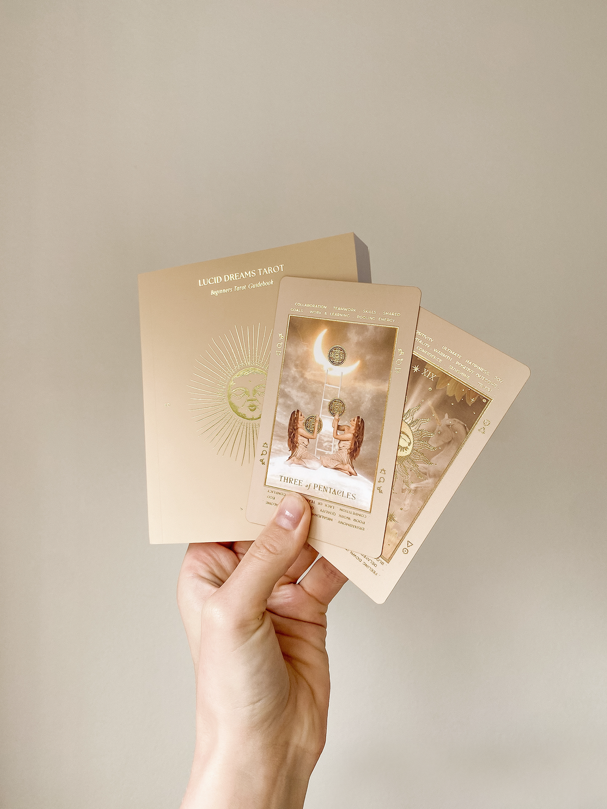 A hand holding three tarot cards and their matching card backs against a plain beige background. The cards feature warm beige, gold, and brown tones with celestial and angelic illustrations. Lucid Dreams Beginner's Tarot Beige Edition IV by ST. SOLEIL with gold foil cards, matte gold edges and guidebook
