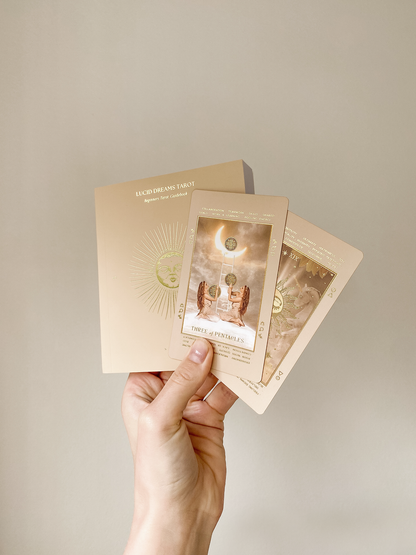 A hand holding three tarot cards and their matching card backs against a plain beige background. The cards feature warm beige, gold, and brown tones with celestial and angelic illustrations. Lucid Dreams Beginner's Tarot Beige Edition IV by ST. SOLEIL with gold foil cards, matte gold edges and guidebook