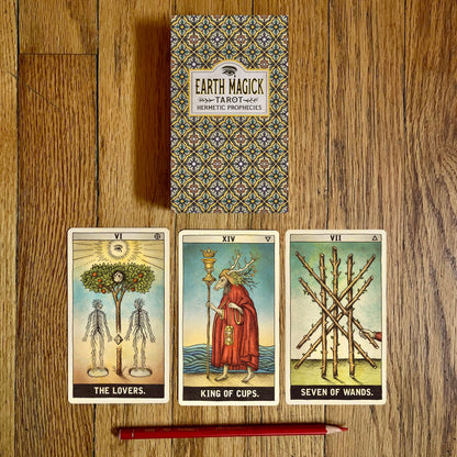 Earth Magick Hex Tarot Deck box and three illustrated tarot cards laid out on a wooden surface: The Lovers, King of Cups, and Seven of Wands, with a red pencil in front.
