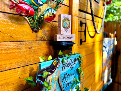 A decorative wooden wall display with colorful metal fish, a small sign reading 'Welcome to Mermaid Cove,' and stacked vintage-style boxes on a black shelf against a warm wood plank background.