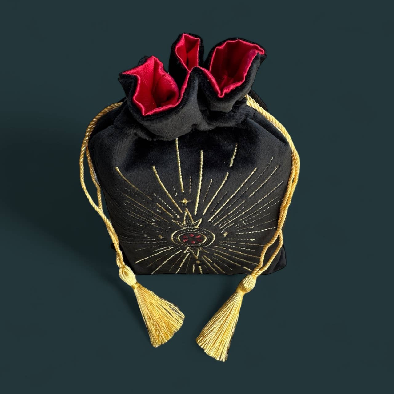 A black velvet drawstring pouch with gold celestial embroidery, a red velvet lining, and gold tassels, set against a dark teal background.