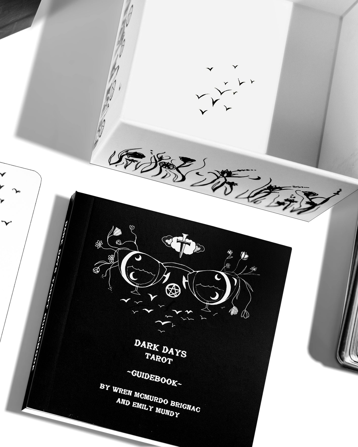 Black tarot guidebook with decorative design on a white surface Dark Days Tarot by Wren McMurdo Brignac featuring square black-and-white tarot cards inspired by the lunar cycle with a 180-page guidebook and collector's box.