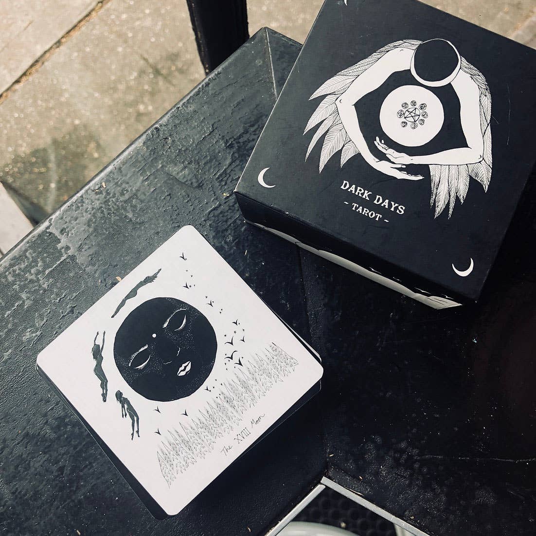 Dark Days Tarot cards on a black surface with a card displayed. Dark Days Tarot by Wren McMurdo Brignac featuring square black-and-white tarot cards inspired by the lunar cycle with a 180-page guidebook and collector's box.