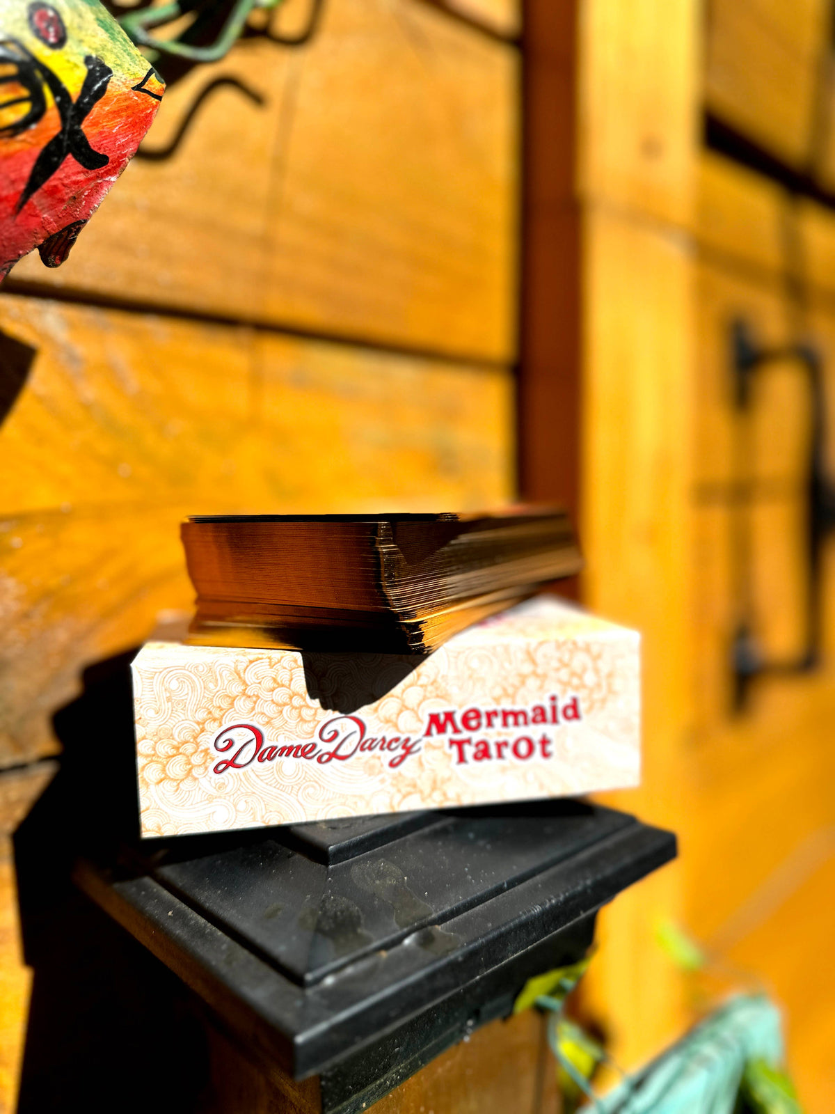 A boxed deck of tarot cards labeled 'Dame Darcy Mermaid Tarot' sits on a black pedestal against a warm wooden background. Gold-gilded card edges on the Dame Darcy Mermaid Tarot Ultra Gold Edition