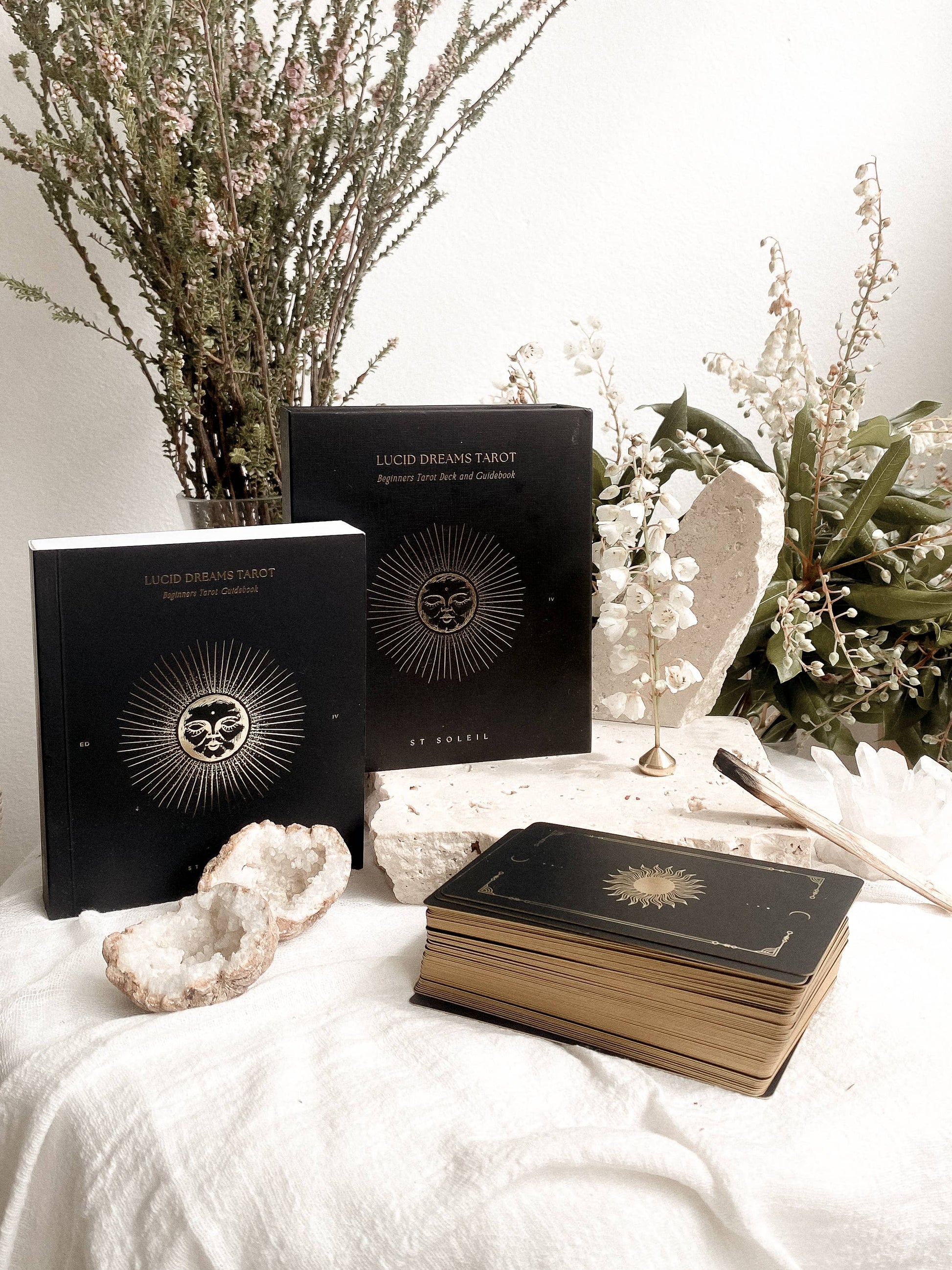 Two black tarot card decks and a matching deck resting on a white fabric surface, styled with dried botanicals, white stones, and a small gold stand. The book covers show a gold sunburst emblem with a central face motif and the title 'LUCKY DREAMS TAROT' and 'BY ROSE'.