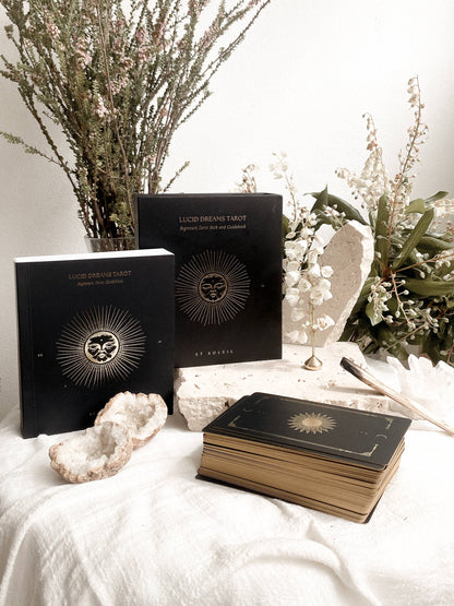 Two black tarot card decks and a matching deck resting on a white fabric surface, styled with dried botanicals, white stones, and a small gold stand. The book covers show a gold sunburst emblem with a central face motif and the title 'LUCKY DREAMS TAROT' and 'BY ROSE'.