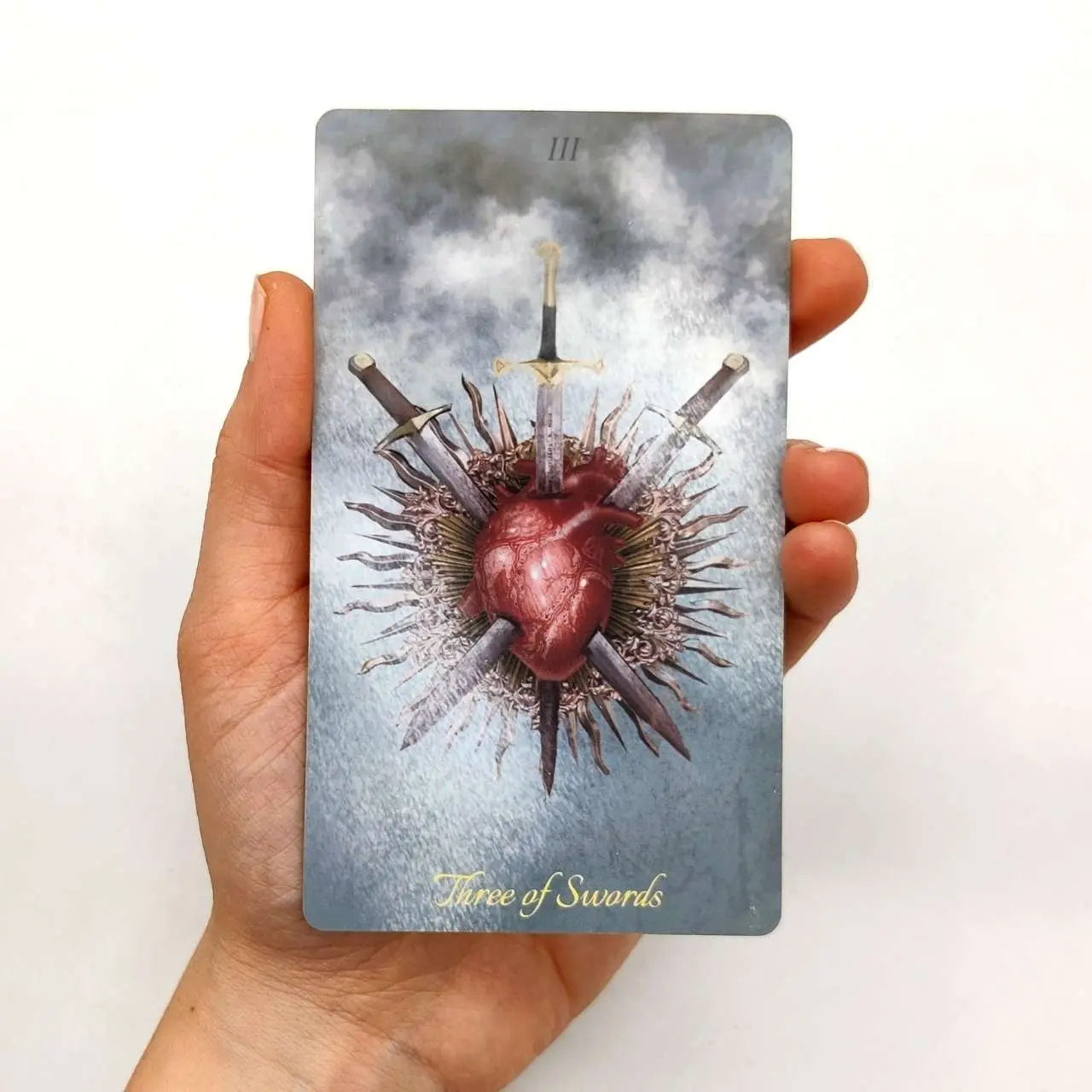 Card with a heart pierced by swords held in a hand against a white background. Three of swords tarot card from the Baroque tarot La Muci 
