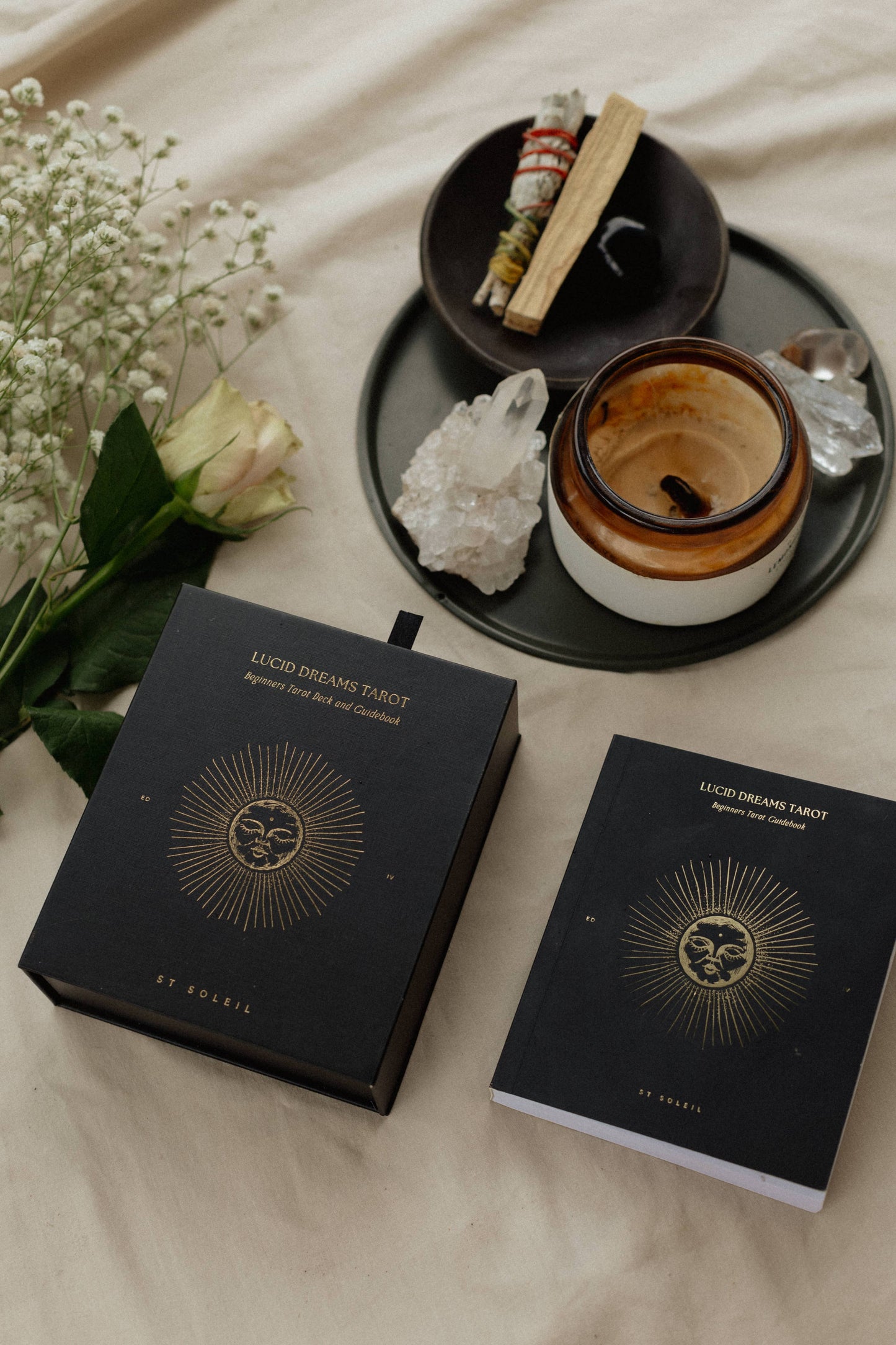 A tarot deck set displayed on a beige fabric background, featuring a black gift box and a black notebook with gold celestial sunburst designs and the text 'LUCKY DREAMING TAROT' and 'by enoria'. A matching white card is partially visible underneath the notebook.
