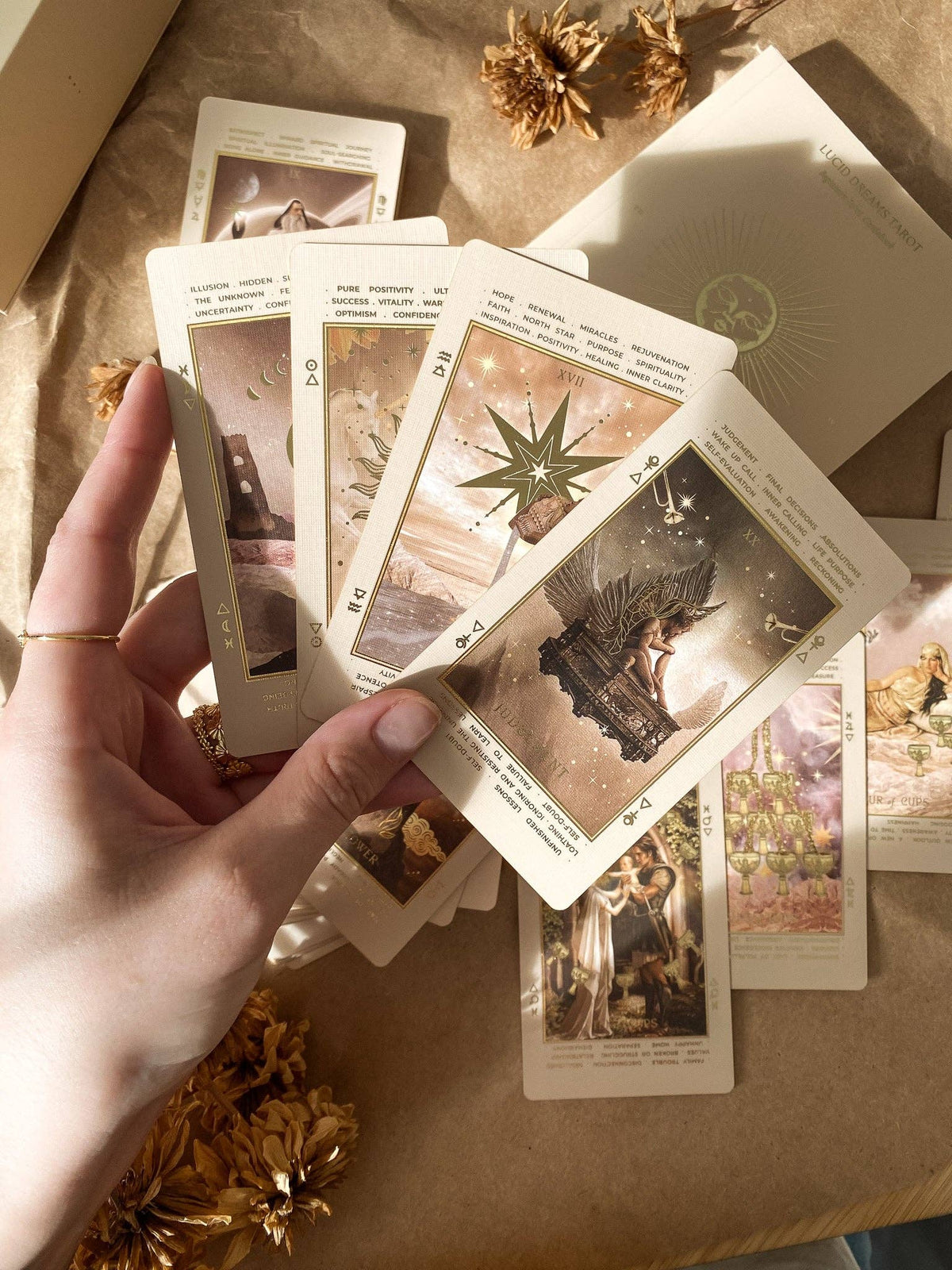 A hand holds a fan of illustrated oracle or tarot-style cards over a brown kraft-paper surface with dried flowers and a cream card envelope or box nearby. Lucid Dreams Beginner's Tarot Beige Edition IV by ST. SOLEIL with gold foil cards, matte gold edges and guidebook