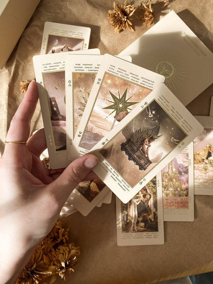 A hand holds a fan of illustrated oracle or tarot-style cards over a brown kraft-paper surface with dried flowers and a cream card envelope or box nearby. Lucid Dreams Beginner's Tarot Beige Edition IV by ST. SOLEIL with gold foil cards, matte gold edges and guidebook