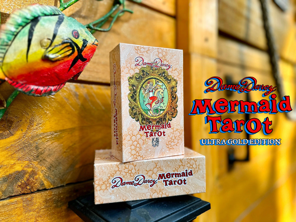 Two decks of Dame Darcy Mermaid Tarot 'ULTRAGOLD EDITION' cards stacked on a black display stand against a rustic wooden background with a decorative fish mask.