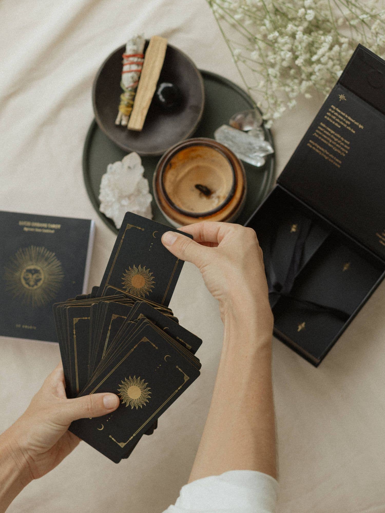 A person's hands holding a fanned deck of black tarot cards with gold celestial designs, with an open black gift box and a tarot card book nearby on a beige fabric surface.