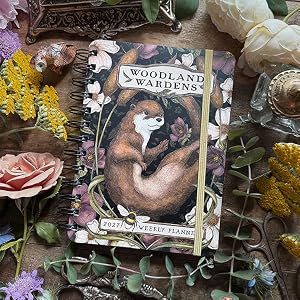 A decorative spiral-bound journal titled 'Woodland Wardens' featuring an illustrated brown bear on the cover, styled in a flat lay with flowers, greenery, and rustic props on a wooden surface.