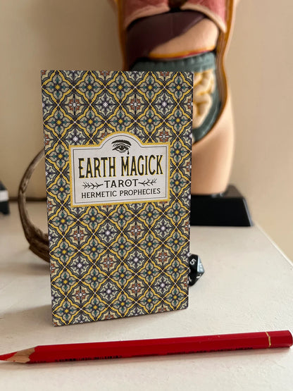 Earth Magick Tarot deck box standing upright on a tabletop, with a red pencil in front and a blurred decorative bust in the background.