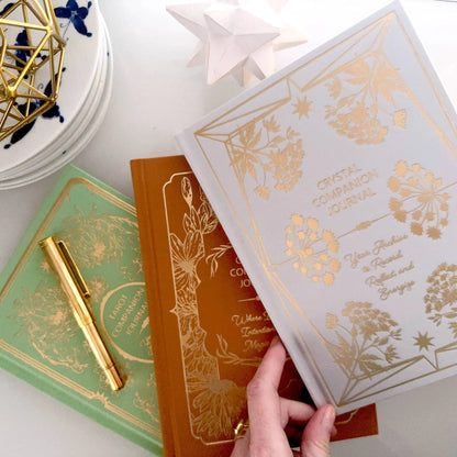 Three books with gold embossed designs on a white surface, one being held open. Tarot Journal Bouchette and Co Oneness Emporium