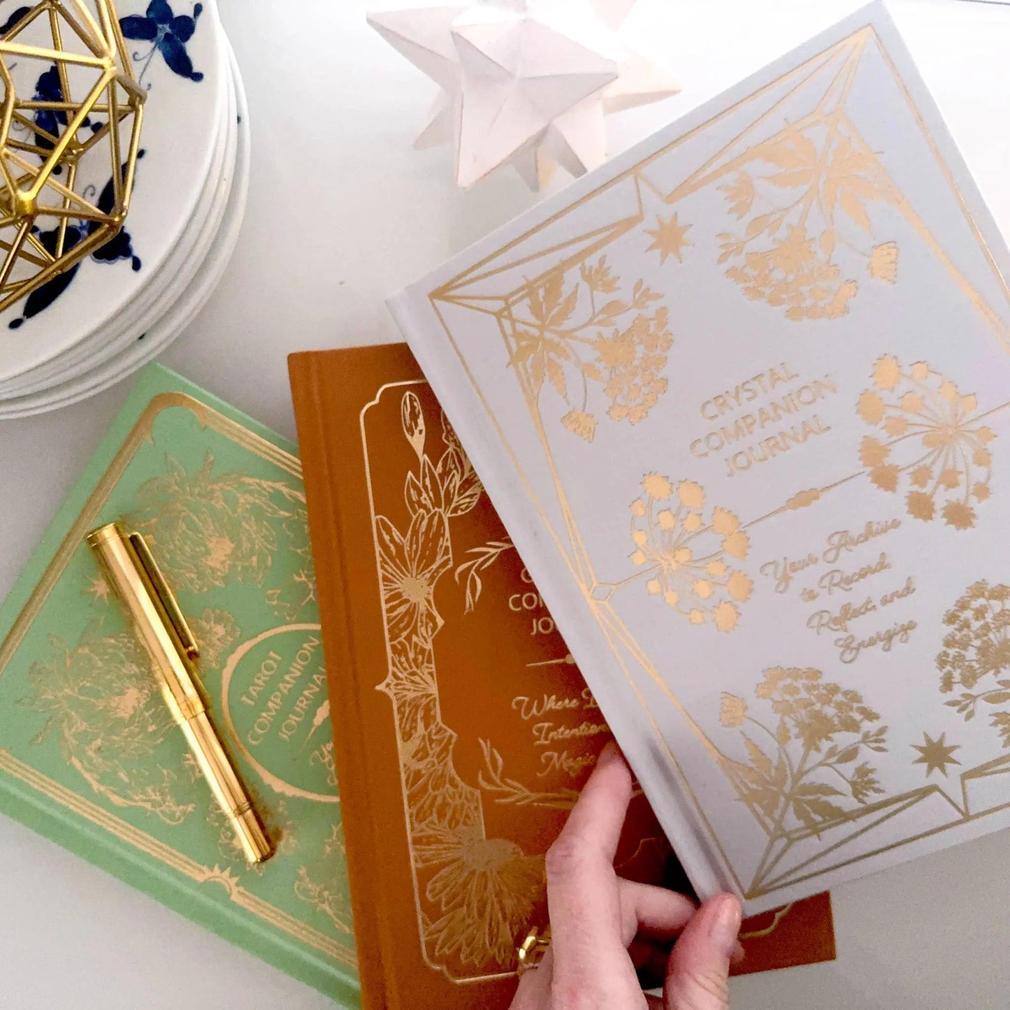 Three books with gold embossed designs on a white surface, one being held open. Crystal companion journal by Bouchette and Co available from Oneness Emporium