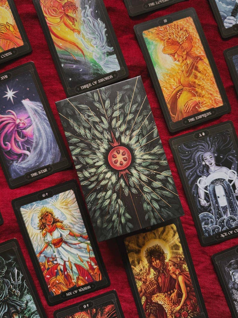 A tarot card deck box sits on a red fabric surface, surrounded by several illustrated tarot cards. The box features a dark green leafy design with a red circular emblem in the center. Asphodelon Tarot by Asphodelon Studio featuring hand-painted Greek mythology artwork, gilded edges, premium matte cards and a deluxe gold foil presentation box.