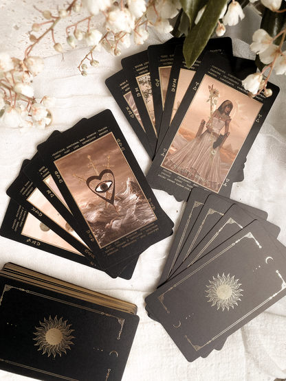 A tarot card deck arranged on a white fabric surface with decorative white flowers. Several cards are fanned out to show illustrated oracle-style cards, and the card backs are visible, including one with a gold sun emblem.