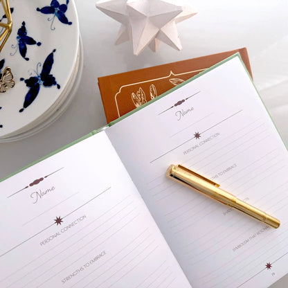 Notebook with a gold pen on a white surface with decorative items. Tarot Journal Bouchette and Co Oneness Emporium