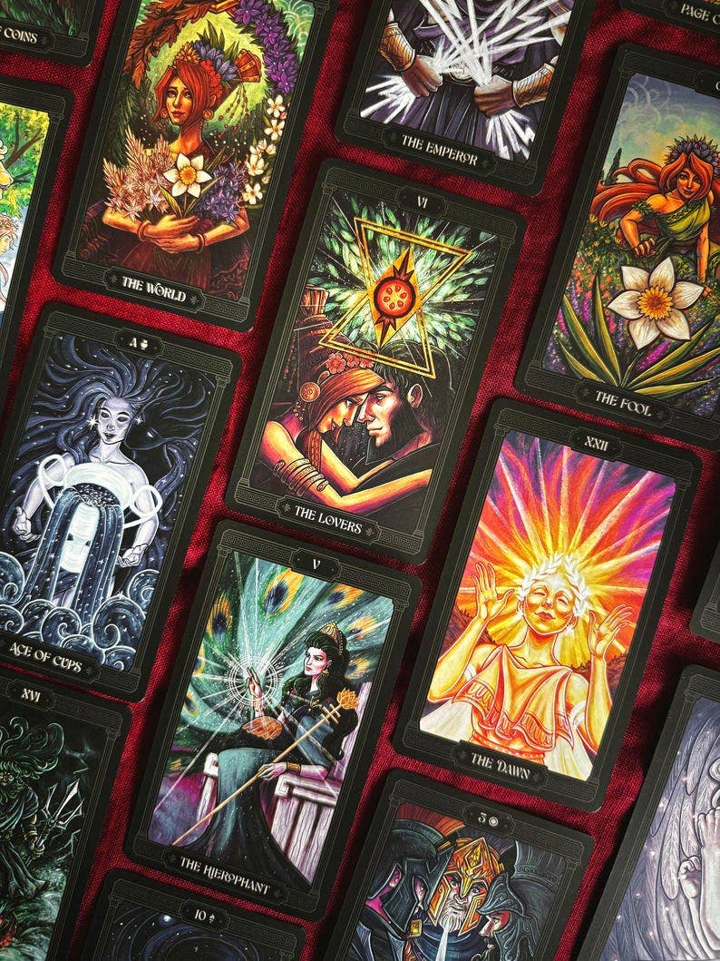 A close-up arrangement of illustrated tarot cards laid over a dark red fabric background. The cards feature colorful fantasy and spiritual artwork with black borders, and several card titles are partially visible. Asphodelon Tarot by Asphodelon Studio featuring hand-painted Greek mythology artwork, gilded edges, premium matte cards and a deluxe gold foil presentation box.