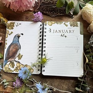 An open spiral-bound planner or journal spread on a rustic wooden surface, showing a January calendar page with lined writing sections and a decorative bird illustration on the left page. The scene is styled with flowers, greenery, and vintage decor in the background.
